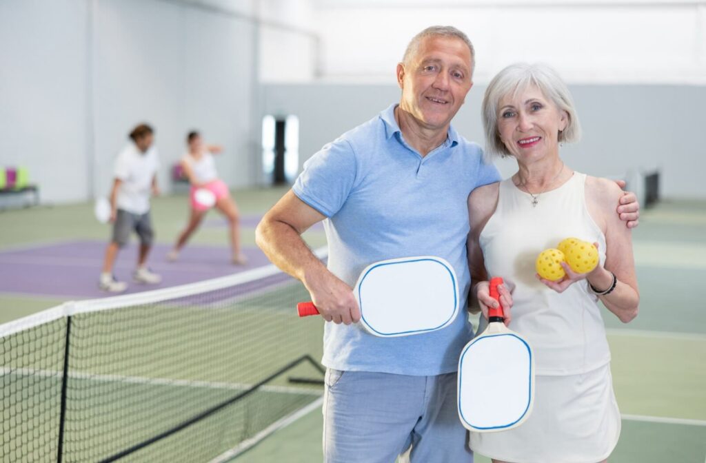 An older couple standing next to each other on a pickleball court holding the right paddles and balls to play pickleball.