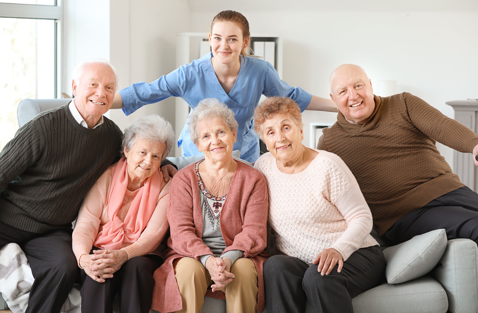 A group of 5 residents in senior living smiling and sitting together on a couch with a happy caregiver behind them.