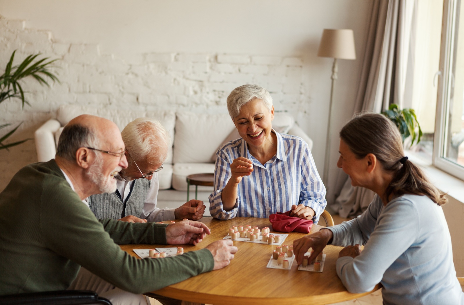 4 older friends in senior living, smiling around a table and laughing over a board game together.
