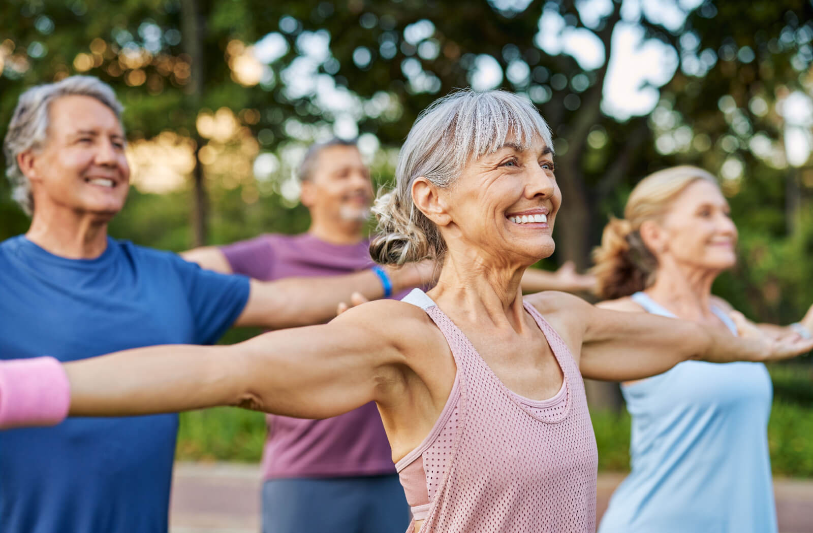 A group of older adults smile while standing with their arms stretched wide during an outdoor exercise class on the lawn in senior living