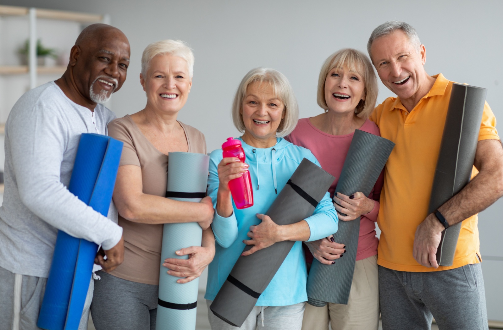 A group of 5 older adults stand together and laugh while holding yoga mats after a group exercise class in senior living