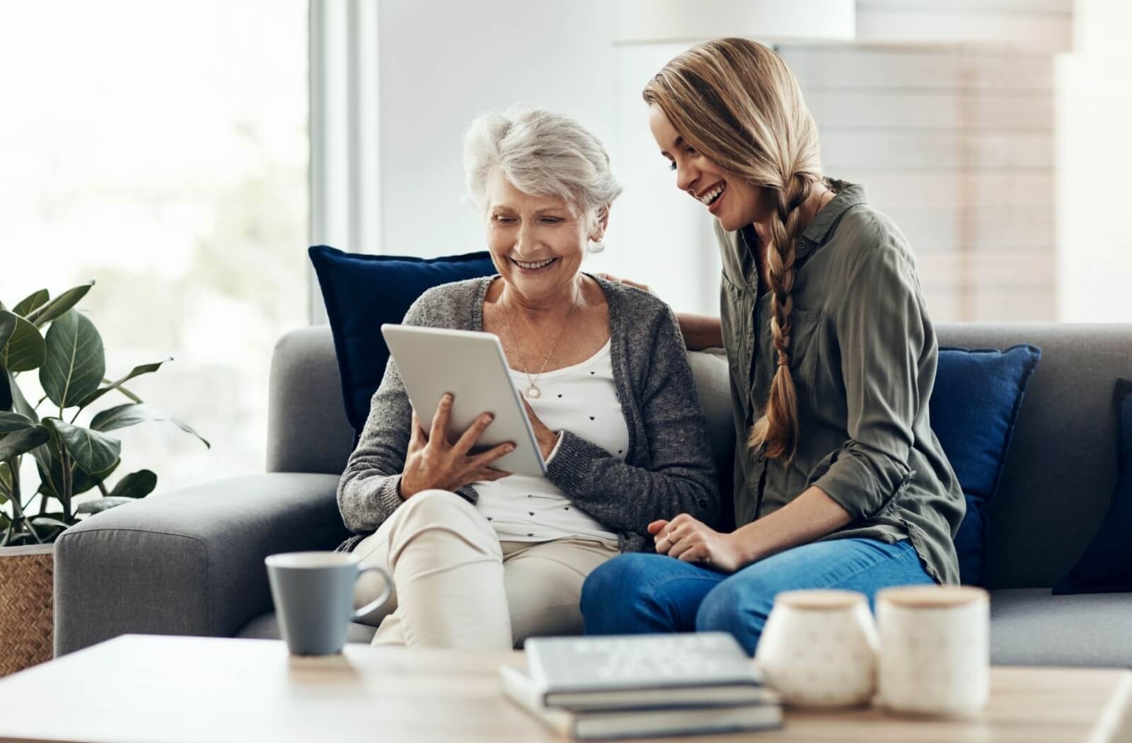 An older adult and their grandchild sit on a couch and watch a video on a digital tablet.
