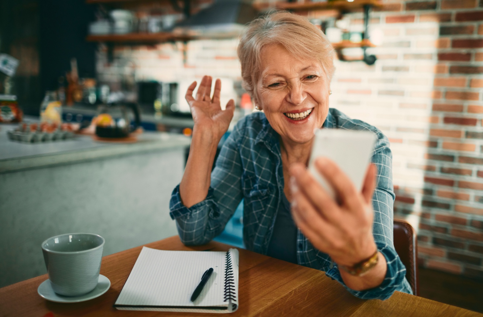 An older adult smiles and waves while video-calling somebody on their cell phone in their kitchen