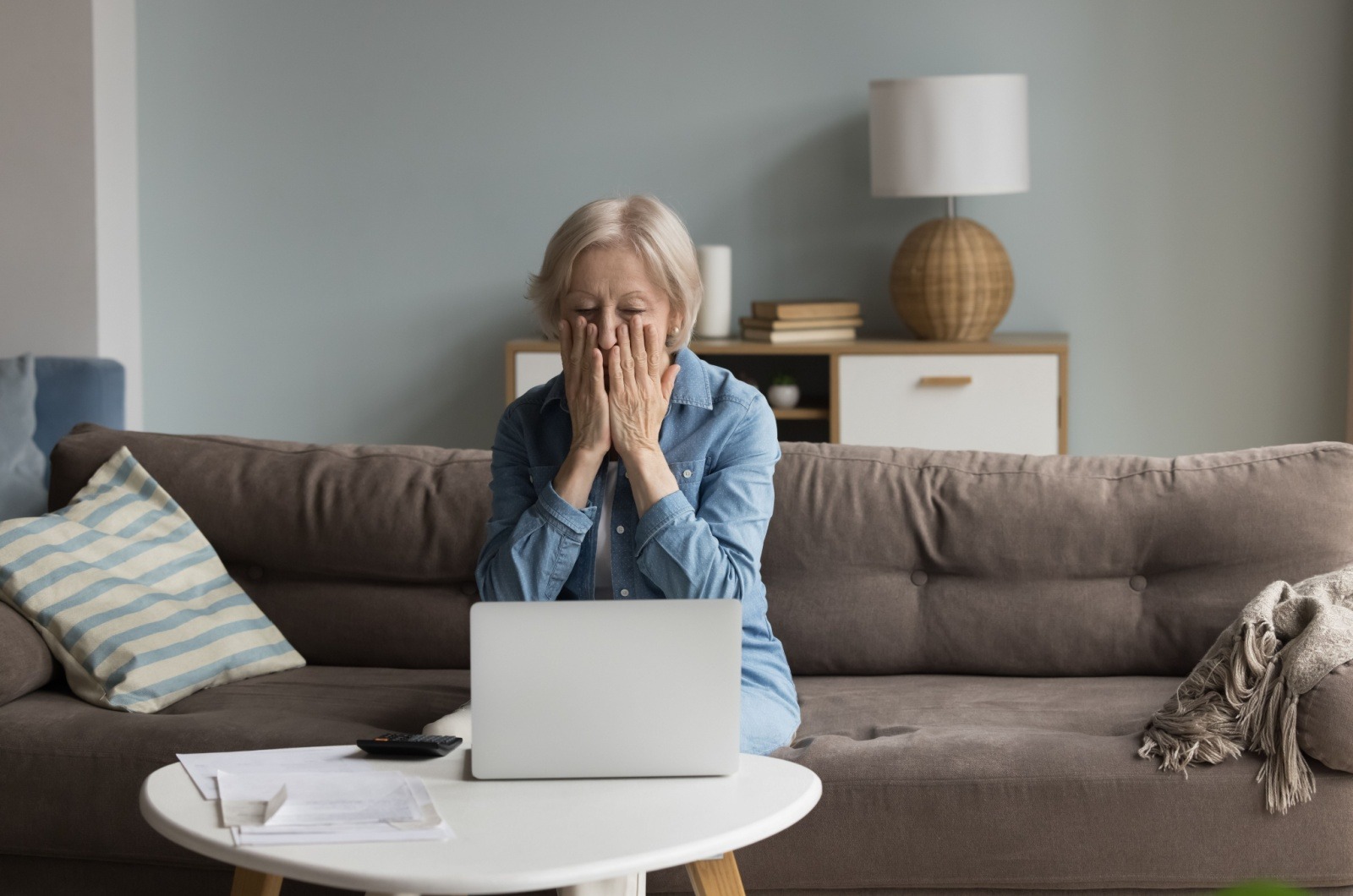 A senior looks shocked and worried while using a laptop and sitting on a brown sofa.