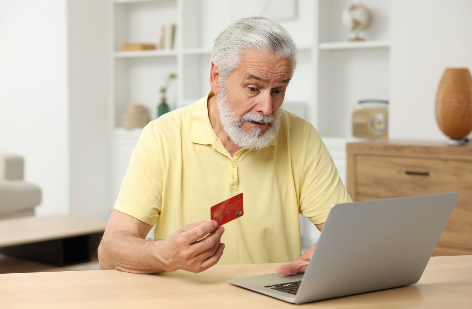 A senior looks concerned while browsing the web on a laptop and holding their credit card in their hand
