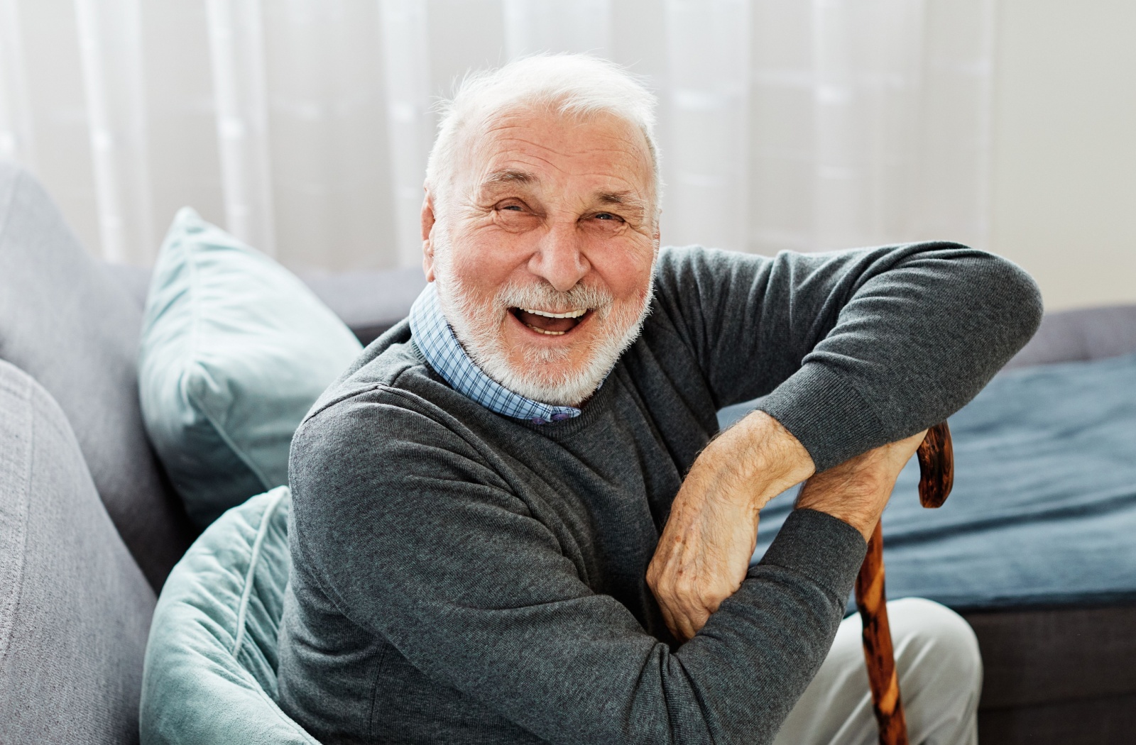 A smiling older adult sitting on a couch with a walking cane in one hand.