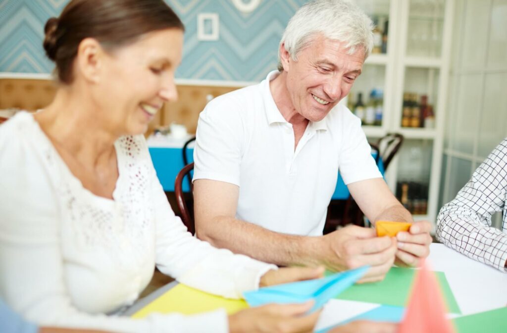 Two smiling older adults sitting at a table creating crafts with colored paper.