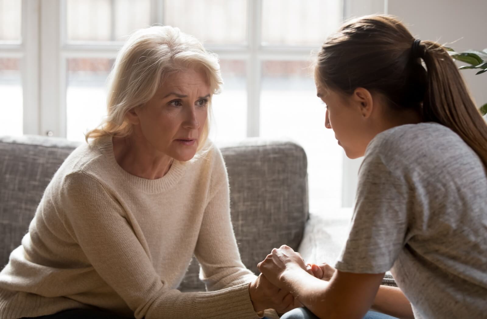 A older woman and her adult daughter sitting on the couch holding hands and having a serious conversation.