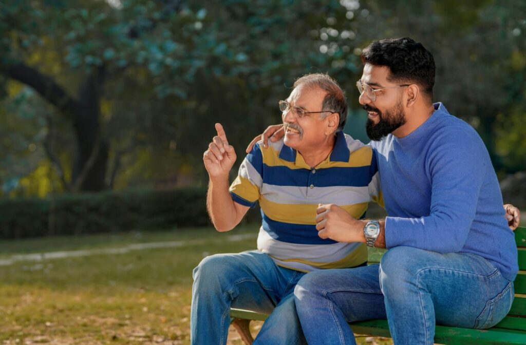 An older man and his adult son sitting on a park bench and smiling as the dad points at something.
