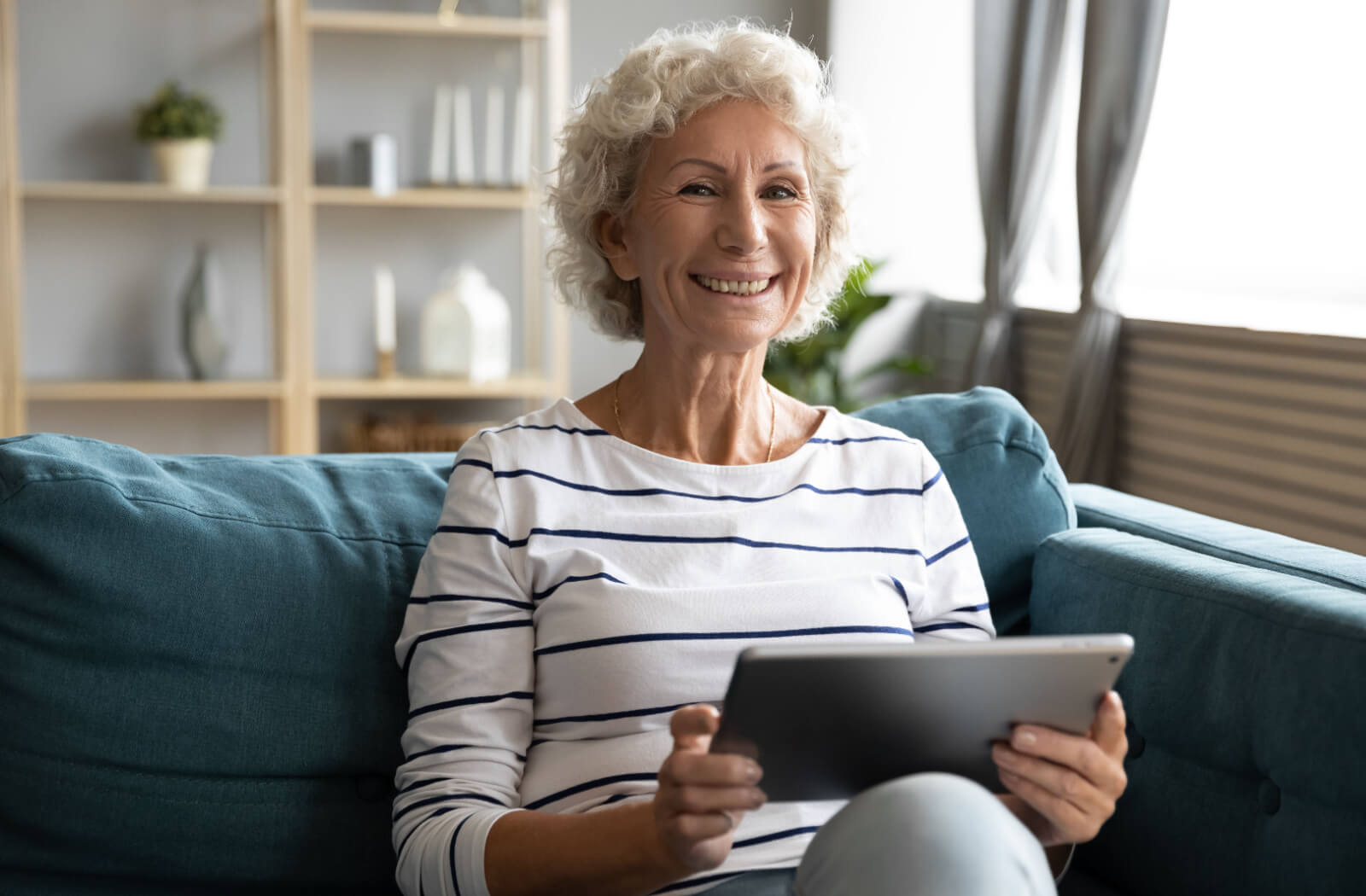 An older adult sitting on the couch in assisted living, smiling and holding an iPad while looking for new free games.