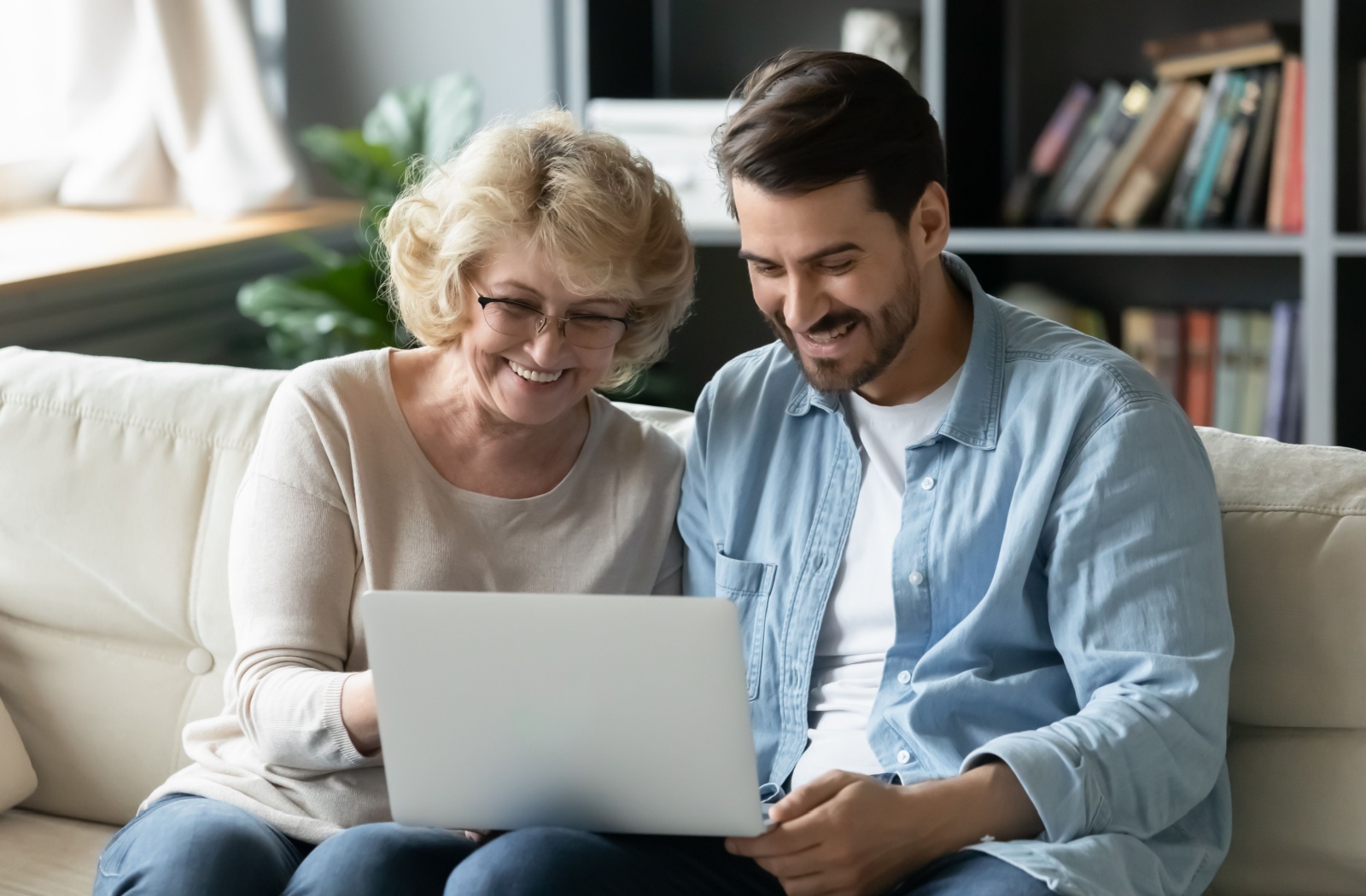 An adult child on the couch beside their senior parent, smiling while they research assisted living communities together.