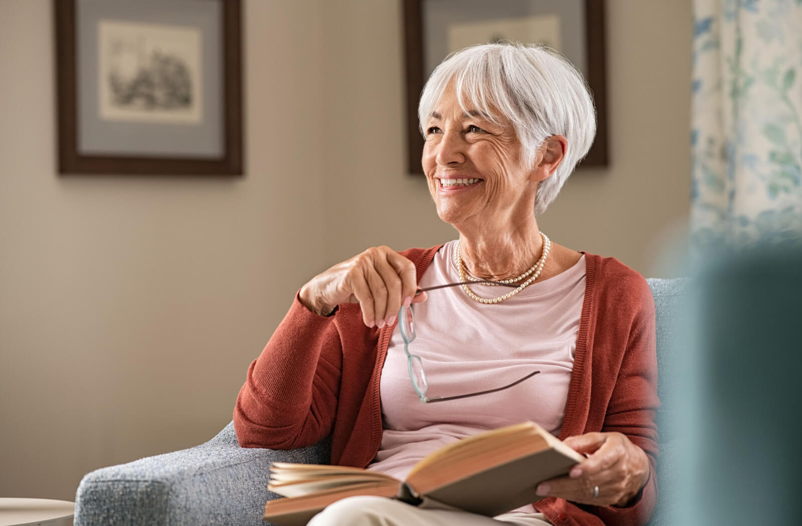 An older adult taking off their glasses to talk to someone out of frame while reading a book in a small armchair.