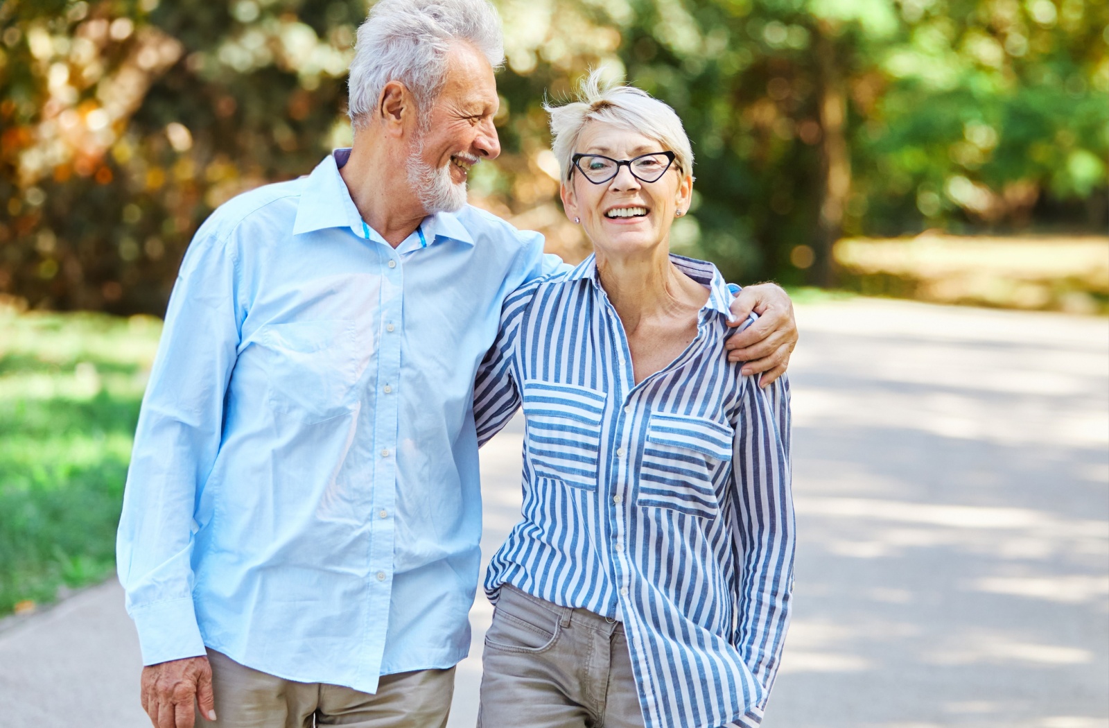 A happy older married couple with arms around one another walking outdoors through the park.