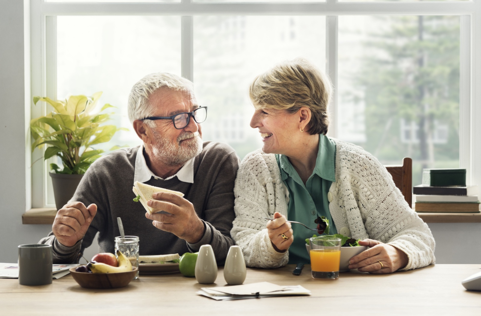 An older married couple smiling at one another while eating lunch in a sunlit room.
